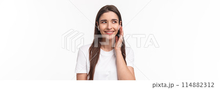 Close-up portrait of attractive young caucasian female in t-shirt, listening favorite song, using wireless earphones, smiling and look away dreamy, touching headphone to change song in playlist 114882312