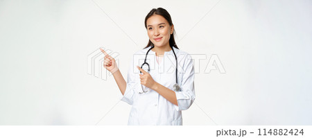 Pleasant smiling female doctor in uniform, pointing fingers and looking left with caring, friendly smile, standing over white background 114882424