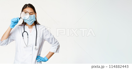 Asian female doctor looking through magnifying glass, examine patient, searching disease, wearing medical mask and rubber gloves, white background Asian female doctor looking through magnifying glass, examine patient, searching disease, wearing medical mask and rubber gloves, white background 114882443
