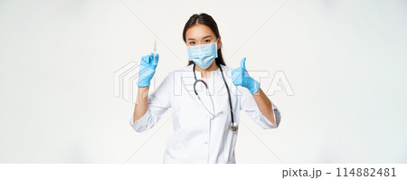 Covid-19 vaccination and healthcare. Asian woman doctor in medical face mask and gloves, holding vaccine syringe and show thumb up, white background Covid-19 vaccination and healthcare. Asian woman doctor in medical face mask and gloves, holding vaccine syringe and show thumb up, white background 114882481