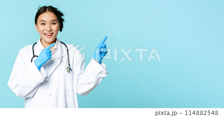 Happy smiling nurse, asian female doctor pointing right, showing promo, clinic advertisement, standing in medical uniform over blue background Happy smiling nurse, asian female doctor pointing right, showing promo, clinic advertisement, standing in medical uniform over blue background 114882548