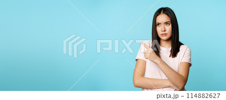 Angry asian woman sulking, frowning upset, pointing fingers at upper left corner, standing in t-shirt over blue background 114882627