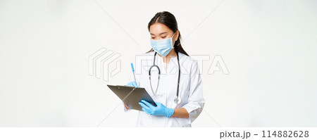 Female asian physician writing down patient info on clipboard, write prescription or diagnose, standing in medical mask with rubber gloves, white background 114882628