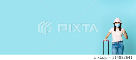 Vertical shot of vaccinated asian girl in medical face mask, going on vacation, standing with suitcase and showing thumb up, blue background 114882641