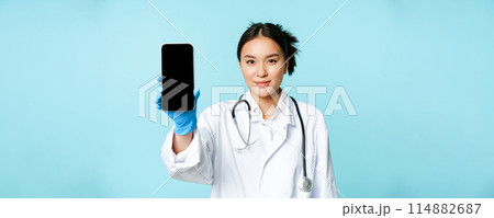 Web medical doctor concept. Smiling asian physician showing mobile phone screen, standing in clinic uniform, blue background Web medical doctor concept. Smiling asian physician showing mobile phone screen, standing in clinic uniform, blue background 114882687