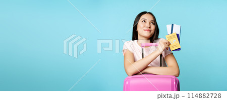 Happy asian female tourist dreaming of trip, lean on suitcase, holding covid-19 international vaccination certificate, two tickets and passport, thinking of journey, blue background 114882728