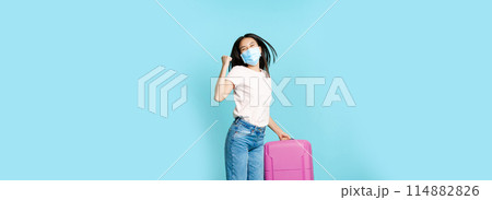 Full length of happy asia female tourist, dancing and jumping with suitcase in medical face mask, excited about vacation, standing over blue background 114882826
