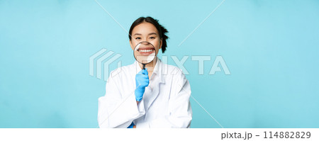 Funny asian female physician, doctor showing her smile with magnifying glass, standing in medical uniform over blue background Funny asian female physician, doctor showing her smile with magnifying glass, standing in medical uniform over blue background 114882829