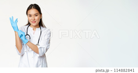 Smiling confident asian female doctor put on medical gloves, getting ready for patient examination, standing in uniform over white background 114882841