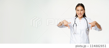 Smiling asian woman doctor, pointing fingers down and showing promo deal, discounts on clinic service, hospital advertisement, white background 114882851
