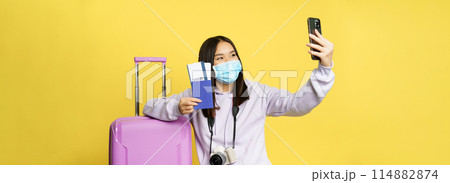 Happy asian woman taking selfie with passport and tickets near suitcase, photographing on smartphone in medical face mask, yellow background Happy asian woman taking selfie with passport and tickets near suitcase, photographing on smartphone in medical face mask, yellow background 114882874