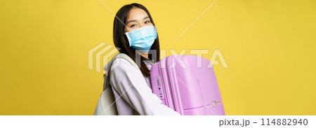 Close up of happy asian girl tourist in medical face mask, holding pink cute suitcase, going on vacation, travelling during pandemic, yellow background Close up of happy asian girl tourist in medical face mask, holding pink cute suitcase, going on vacation, travelling during pandemic, yellow background 114882940