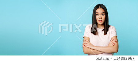 Portrait of young asian woman listening with confused, shocked face expression, standing in t-shirt over blue background 114882967