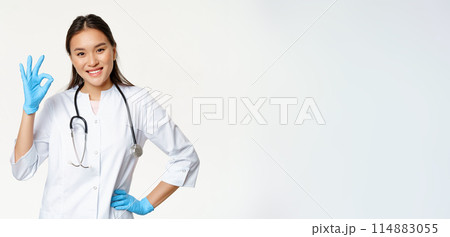 Smiling asian female physician, nurse in rubber gloves and medical uniform shows okay sign, approves and likes smth good, standing over white background 114883055