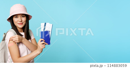 Happy asian girl tourist got vaccinated to travel abroad, showing arm with plaster and passport with two tickets, booking tour, travelling during pandemic, blue background 114883059
