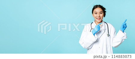Happy smiling nurse, asian female doctor pointing right, showing promo, clinic advertisement, standing in medical uniform over blue background Happy smiling nurse, asian female doctor pointing right, showing promo, clinic advertisement, standing in medical uniform over blue background 114883073