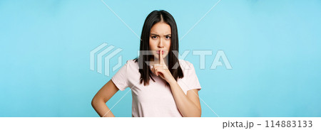 Angry asian woman shushing, frowning in disapproval and hushing, taboo sign, silence gesture, standing over blue background 114883133