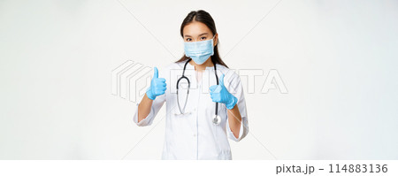 Covid-19 and medical workers concept. Smiling asian woman doctor in face mask and rubber gloves shows thumbs up, white background Covid-19 and medical workers concept. Smiling asian woman doctor in face mask and rubber gloves shows thumbs up, white background 114883136