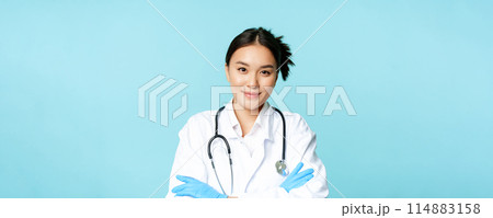 Smiling asian woman doctor, nurse in gloves and clinic uniform, cross arms and looking determined, blue background Smiling asian woman doctor, nurse in gloves and clinic uniform, cross arms and looking determined, blue background 114883158