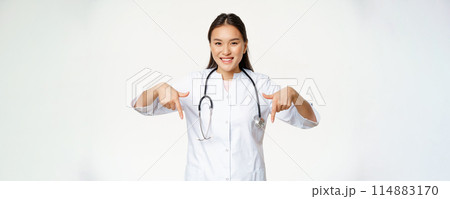 Smiling asian woman doctor, pointing fingers down and showing promo deal, discounts on clinic service, hospital advertisement, white background Smiling asian woman doctor, pointing fingers down and showing promo deal, discounts on clinic service, hospital advertisement, white background 114883170