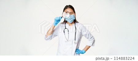 Asian female doctor looking through magnifying glass, examine patient, searching disease, wearing medical mask and rubber gloves, white background 114883224
