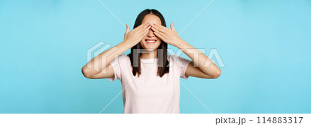 Happy smiling asian girl covers ears and waits for surprise, standing in t-shirt over blue background 114883317