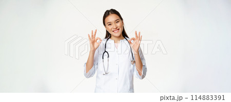 Female smiling doctor, asian healthcare worker in medical clinic uniform shows okay signs, approve smth, give patient permission, stands over white background 114883391