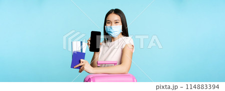 Tourism and healthcare concept. Asian female tourist with suitcare, wearing medical face mask for flight, showing smartphone screen and passport with tickets, vaccination certificate 114883394