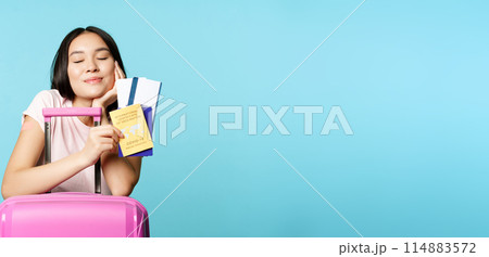 Smiling dreamy asian tourist, girl thinks of travelling, holding passport and tickets, coronavirus international vaccination certificate, going on trip abroad, blue background 114883572