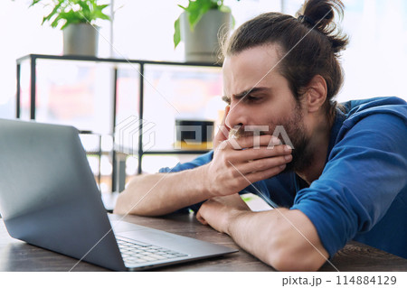 Young tired sleepy man looking at laptop, yawning covering his mouth with hand Young tired sleepy man looking at laptop, yawning covering his mouth with hand 114884129