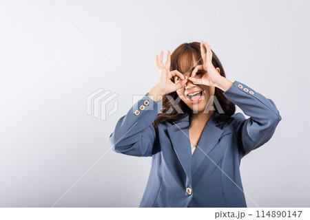 A cheerful young Asian woman stands gracefully, forming an 'OK' hand symbol near her eye, signifying agreement and positivity, in a studio with copy space. 114890147