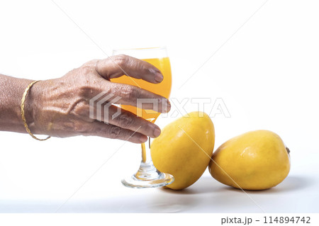 Senior woman hand holding a glass of mango juice next to ripe two mangoes on a white background. 114894742
