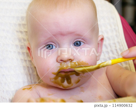 Mom feeding little boy with broccoli puree. Child at the age of six months eats broccoli while sitting on a baby chair. 114900632