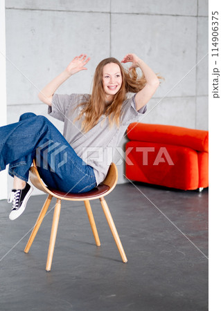 Beautiful woman with braces in jeans and a gray T-shirt sits relaxed on a chair against a gray wall and smiles 114906375