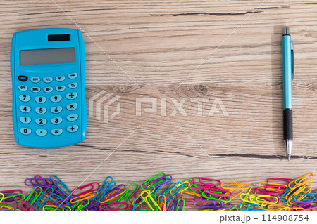 Colorful office paper clips. Electronic calculator. Blue pen. Wooden desk top. Colorful office paper clips. Electronic calculator. Blue pen. Wooden desk top. 114908754