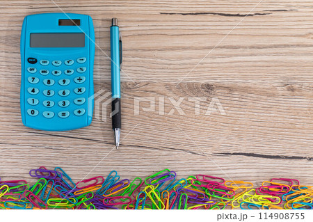 Colorful office paper clips. Electronic calculator. Blue pen. Wooden desk top. Colorful office paper clips. Electronic calculator. Blue pen. Wooden desk top. 114908755