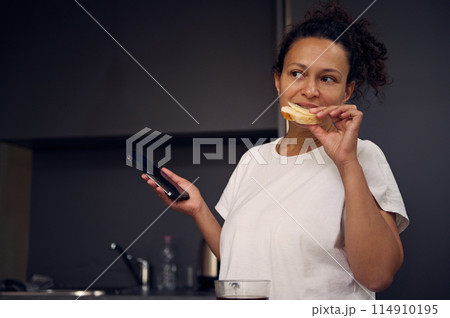 Authentic young adult woman 40s, eating sandwich with cheese and holding her smartphone in hands, smiling looking aside, standing at kitchen counter in minimalist home kitchen interior. Authentic young adult woman 40s, eating sandwich with cheese and holding her smartphone in hands, smiling looking aside, standing at kitchen counter in minimalist home kitchen interior. 114910195