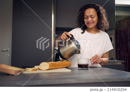 Smiling multi ethnic woman at home, making coffee. Young pretty brunette n white pajamas, standing at kitchen table, preparing breakfast, poring water from a teapot into a glass and making sandwiches 114910204