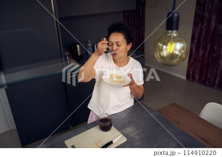 Latin American young woman 40s, smiling, drinking coffee and eating healthy muesli for breakfast in the morning at home kitchen, dressed in white pajamas 114910220