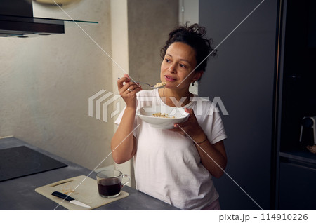 Young pretty woman in white t-shirt, eating a bowl of muesli at home. People. Food consumerism. Diet and healthy lifestyle Young pretty woman in white t-shirt, eating a bowl of muesli at home. People. Food consumerism. Diet and healthy lifestyle 114910226