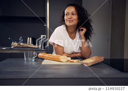 Authentic curly haired brunette woman in white t-shirt, dreamily looking aside, standing at kitchen countertop with a loaf of fresh bread and slice of cheese for breakfast. People. Lifestyle Authentic curly haired brunette woman in white t-shirt, dreamily looking aside, standing at kitchen countertop with a loaf of fresh bread and slice of cheese for breakfast. People. Lifestyle 114910227