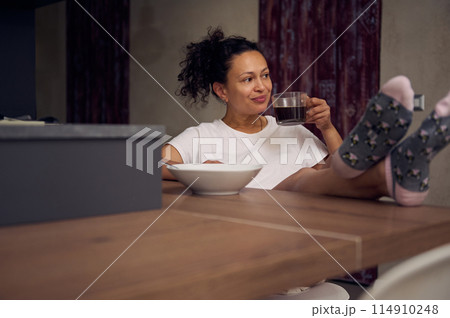 Pretty woman in pajamas, relaxing over cup of coffee. Happy young adult sitting at kitchen table, drinking hot coffee in the morning during her breakfast. Full size portrait Pretty woman in pajamas, relaxing over cup of coffee. Happy young adult sitting at kitchen table, drinking hot coffee in the morning during her breakfast. Full size portrait 114910248