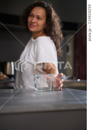 Happy woman taking out a glass cup from a cupboard, putting it on the kitchen counter, smiling looking aside, standing in modern minimalist home kitchen interior. Morning routine. Healthy lifestyle. Happy woman taking out a glass cup from a cupboard, putting it on the kitchen counter, smiling looking aside, standing in modern minimalist home kitchen interior. Morning routine. Healthy lifestyle. 114910250