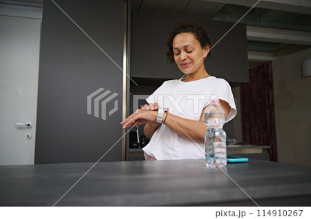 Multi ethnic smiling heathy young woman checking smart wrist watch. A bottle of pure water on the foreground. Fitness app, mobile app. People. Health care and modern wireless technology 114910267