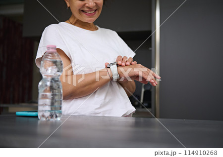 Close-up view of hands of a smiling heathy young woman checking smart wrist watch. A bottle of pure water on the foreground. Fitness app, mobile app. People. Health care and modern wireless technology 114910268