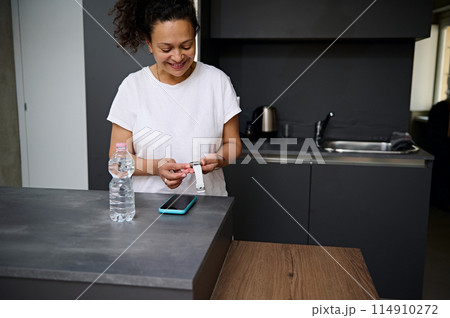 Smiling pretty woman in white t-shirt holding modern white smart wrist watch with black mockup digital screen, standing at kitchen counter in the minimalist home interior. Copy advertising space 114910272