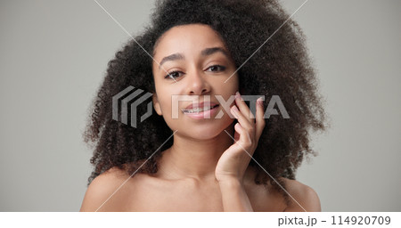 Beauty and healthcare concept - beautiful African American woman with curly afro hairstyle and clean, healthy skin touching face with hand, doing facial massage, posing and looking at camera 114920709