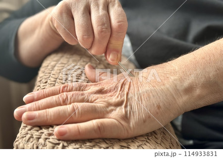 An unknown elderly woman, sitting in an armchair, massages her hand with pressure on the other hand using a silver needle. Health maintenance and Promotion Program. Close-up 114933831