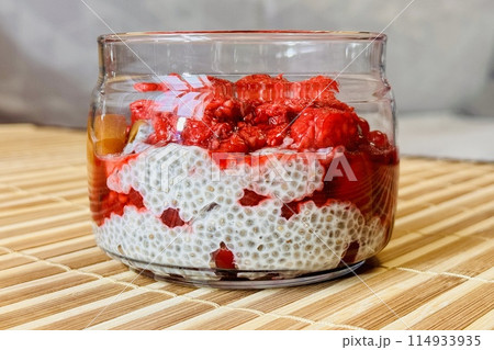 Healthy Chia pudding with coconut milk in glass container in view can with fresh berries as raspberry, red currant, cranberry and pieces of peach on bamboo background on store counter. Close-up Healthy Chia pudding with coconut milk in glass container in view can with fresh berries as raspberry, red currant, cranberry and pieces of peach on bamboo background on store counter. Close-up 114933935