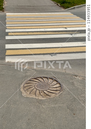A sunny day view of a crosswalk with alternating yellow and white stripes beside a manhole cover A sunny day view of a crosswalk with alternating yellow and white stripes beside a manhole cover 114933951